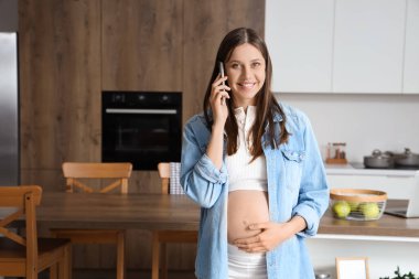 Genç hamile kadın mutfakta cep telefonuyla konuşuyor.