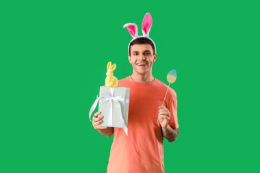 Tavşan kulaklı mutlu genç adam hediye kutusu, oyuncak tavşan ve yeşil arka planda kağıt yumurta. Paskalya kutlaması