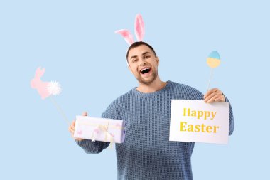 Tavşan kulaklı mutlu genç adam hediye kutusu, tebrik kartı ve mavi arka planda kağıt süslemeler. Paskalya kutlaması