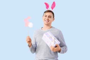 Tavşan kulaklı, hediye kutusu ve mavi arka planda kağıttan tavşanı olan mutlu genç adam. Paskalya kutlaması