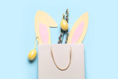 Shopping bag with paper bunny ears, Easter eggs and pussy willow branches on blue background