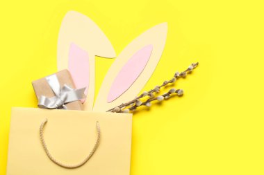 Shopping bag with paper Easter bunny ears, gift box and pussy willow branches on yellow background