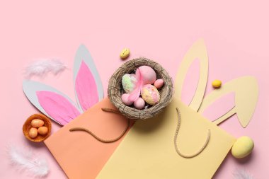 Paskalya tavşanı kulaklı alışveriş torbaları, pembe arka planda yuvalar ve yumurtalar.