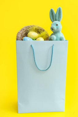 Sarı arka planda oyuncak Paskalya tavşanı, yuva ve yumurta dolu alışveriş torbası.