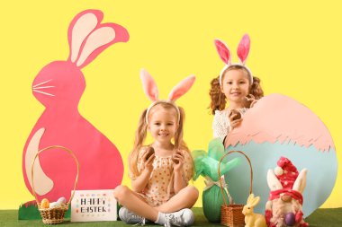 Çikolatalı yumurta ve çimlerin üzerinde sarı arka planda dekoru olan tavşan kulaklı şirin küçük kızlar.