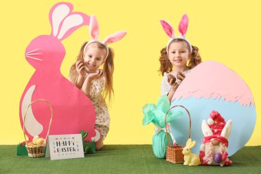 Çikolatalı yumurta ve çimlerin üzerinde sarı arka planda dekoru olan tavşan kulaklı şirin küçük kızlar.