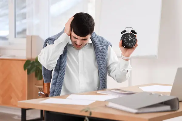 Stressed young businessman with alarm clock in office
