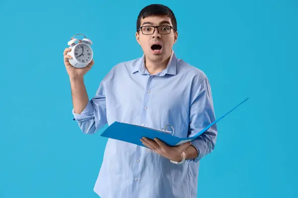Stressed young businessman with alarm clock and folder on blue background