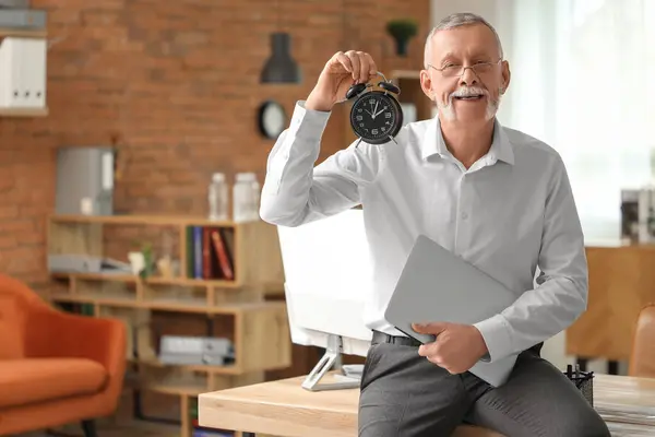Mature businessman with alarm clock and laptop in office