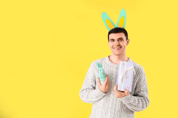 黄色の背景にギフトボックスと玩具ウサギを抱えたバニーの耳の幸せな若い男 イースターのお祝い — ストック写真