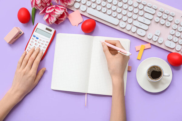 Женские руки с чистым блокнотом, калькулятор, клавиатура и пасхальные яйца на сиреневом фоне