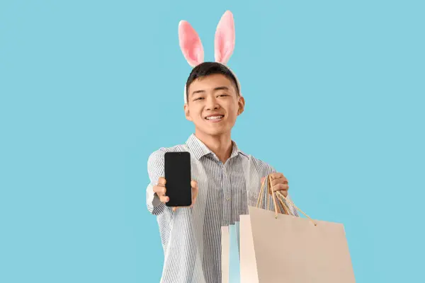 Junger Asiatischer Mann Mit Hasenohren Handy Und Einkaufstaschen Auf Blauem — Stockfoto