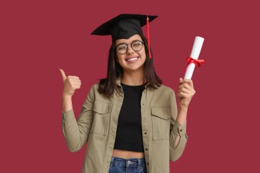 Mezuniyet şapkalı mutlu bayan öğrenci kırmızı arka planda baş parmağını gösteriyor.