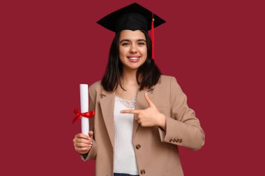 Mezuniyet şapkalı mutlu bayan öğrenci kırmızı arka plandaki diplomayı gösteriyor.