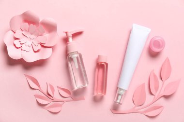 Temizleyici, tonik, yüz kremi, kağıt çiçekler ve pembe arka planda yapraklar.