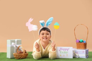 Tavşan kulaklı şirin bir çocuk. Yumurta sepetleri, hediye kutuları ve bej arka planda çimlerin üzerinde duran Paskalya bayramı kartpostallarıyla.