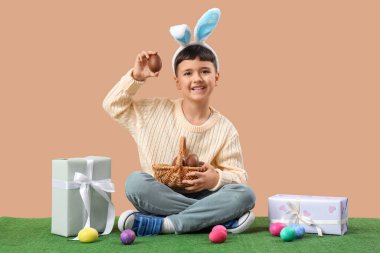 Tavşan kulaklı şirin çocuk. Çikolatalı yumurta sepeti ve paskalya için hediye kutularıyla bej arka planda çimlerin üzerinde oturuyor.