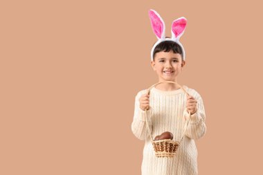 Tavşan kulaklı şirin çocuk bej arka planda Paskalya için çikolatalı yumurta sepeti tutuyor.