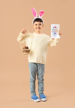 Tavşan kulaklı şirin çocuk çikolata yumurtalı sepet tutuyor ve bej arka planda Paskalya için neşeli kartpostal yolluyor.