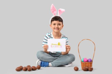 Tavşan kulaklı şirin bir çocuk Paskalya ve sepet için beyaz arka planda çikolatalı yumurtalar tutan neşeli bir kartpostal tutuyor.