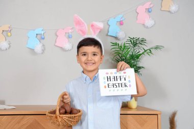 Tavşan kulaklı şirin çocuk elinde çikolata yumurtaları ve Paskalya bayramı kartpostallarıyla sepet tutuyor.