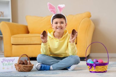 Evde paskalya için sepetli, çikolata yumurtalı ve neşeli kartpostallı tavşan kulaklı şirin bir çocuk.