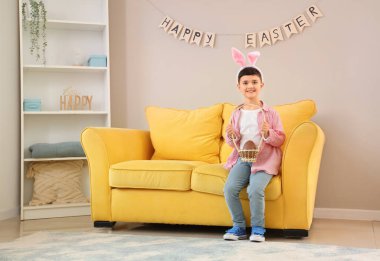 Paskalya için çikolata yumurtası sepeti olan tavşan kulaklı şirin çocuk evde kanepede oturuyor.