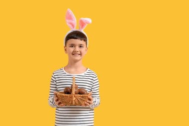 Tavşan kulaklı şirin çocuk, sarı arka planda çikolata yumurtalı sepet tutuyor.