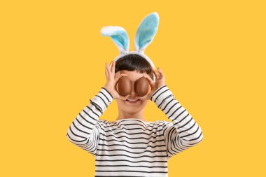 Sarı arka planda Paskalya için çikolatalı yumurtalarla tavşan kulaklı şirin bir çocuk.