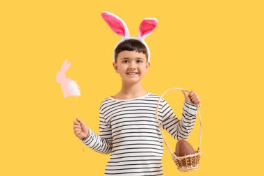 Tavşan kulaklı şirin bir çocuk. Sarı arka planda Paskalya için çikolata yumurtası ve dekor dolu bir sepet tutuyor.
