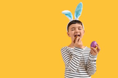 Tavşan kulaklı şirin çocuk, sarı arka planda Paskalya için çikolatalı yumurta yiyor.