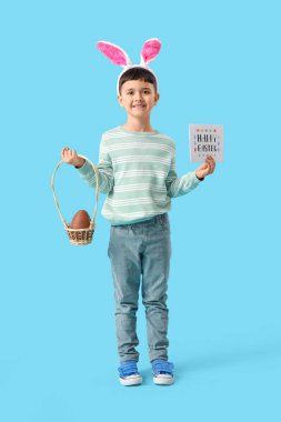 Tavşan kulaklı şirin çocuk elinde çikolata yumurtası sepeti ve mavi arka planda Paskalya bayramı için kartpostal tutuyor.