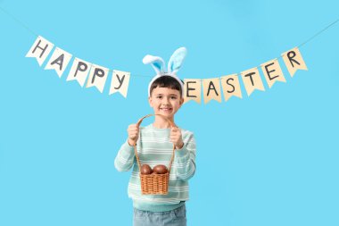 Tavşan kulaklı şirin bir çocuk elinde çikolata yumurtası sepeti ve mavi arka planda Mutlu Paskalyalar yazan bir çelenk tutuyor.