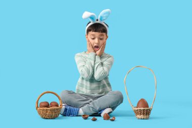 Şok olmuş tavşan kulaklı küçük çocuk mavi arka planda oturan Paskalya için çikolata yumurtası sepetleriyle.