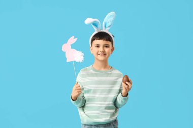 Kafasında çikolata yumurtası ve mavi arka planda Paskalya için dekoru olan tavşan kulaklı şirin bir çocuk.