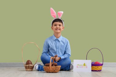 Tavşan kulaklı şirin bir çocuk. Elinde sepetlerce çikolata yumurtası ve Paskalya için yeşil duvarın yanında duran neşeli kartpostal var.