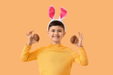 Turuncu arka planda Paskalya için çikolatalı yumurtalarla tavşan kulaklı şirin bir çocuk.