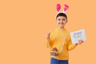 Tavşan kulaklı şirin bir çocuk çikolata yumurtası ve turuncu arka planda Paskalya bayramı kartpostalı taşıyan bir sepet tutuyor.