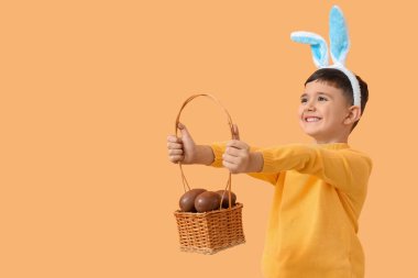 Tavşan kulaklı şirin bir çocuk. Turuncu arka planda Paskalya için çikolatalı yumurta sepeti tutuyor.