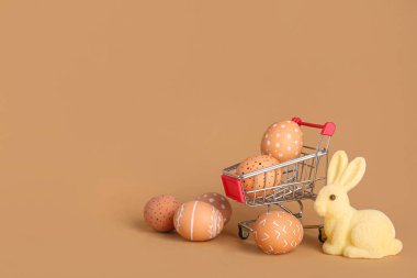 Paskalya yumurtaları ve bej arka planda tavşan heykelciği olan küçük bir alışveriş arabası.