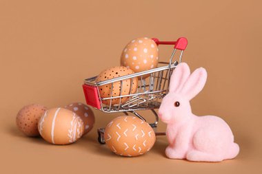 Paskalya yumurtaları ve bej arka planda tavşan heykelciği olan küçük bir alışveriş arabası.