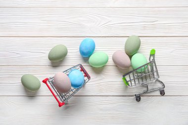 Beyaz ahşap arka planda Paskalya yumurtaları olan küçük alışveriş arabaları.
