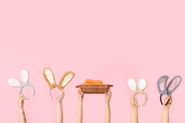 Paskalya tavşanı kulaklıklarını tutan kadın elleri ve pembe arka planda havuç dolu sepet.