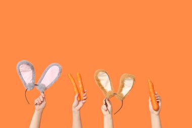 Paskalya tavşanı kulaklıkları ve turuncu arka planda havuç tutan kadın elleri.