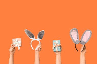Paskalya tavşanı kulaklıkları ve turuncu arka planda hediye kutuları tutan kadın elleri.