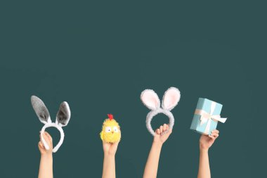 Paskalya tavşanı kulaklığı takan, hediye kutusu ve koyu yeşil arka planda oyuncak tavuk taşıyan kadın elleri.