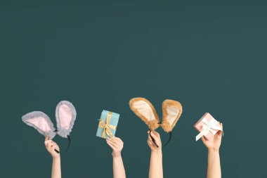 Paskalya tavşanı kulaklıklarını tutan kadın elleri ve koyu yeşil arka planda hediye kutuları.