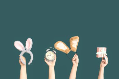 Paskalya tavşanı kulaklığı takan, hediye kutusu ve koyu yeşil arka planda çalar saati olan kadın eller.