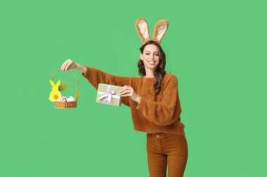 Paskalya tavşanı kulaklıklı mutlu genç kadın elinde çiçekli sepet ve yeşil arka planda hediye kutusu tutuyor.