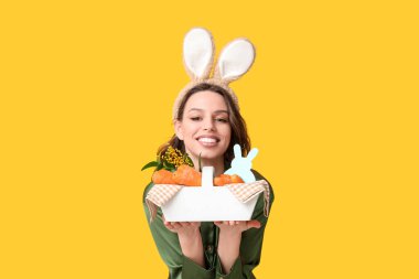 Paskalya tavşanı kulaklıklı mutlu genç kadın sarı arka planda havuçlu sepet tutuyor.